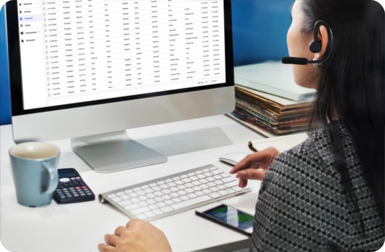Woman with headset at a desk using a keyboard and tablet with a large monitor displaying a payments dashboard and a coffee mug nearby